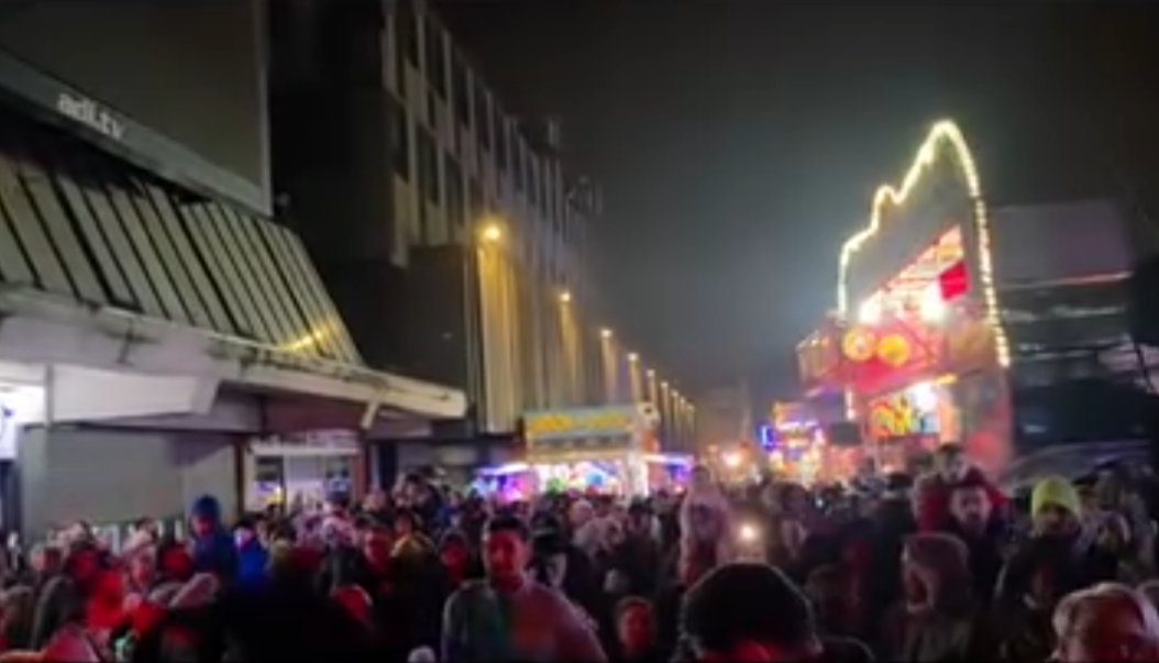 Crowds at Irvine town centre at night during the Christmas event, with fairground rides illuminated in the distance and Christmas lights along the street.