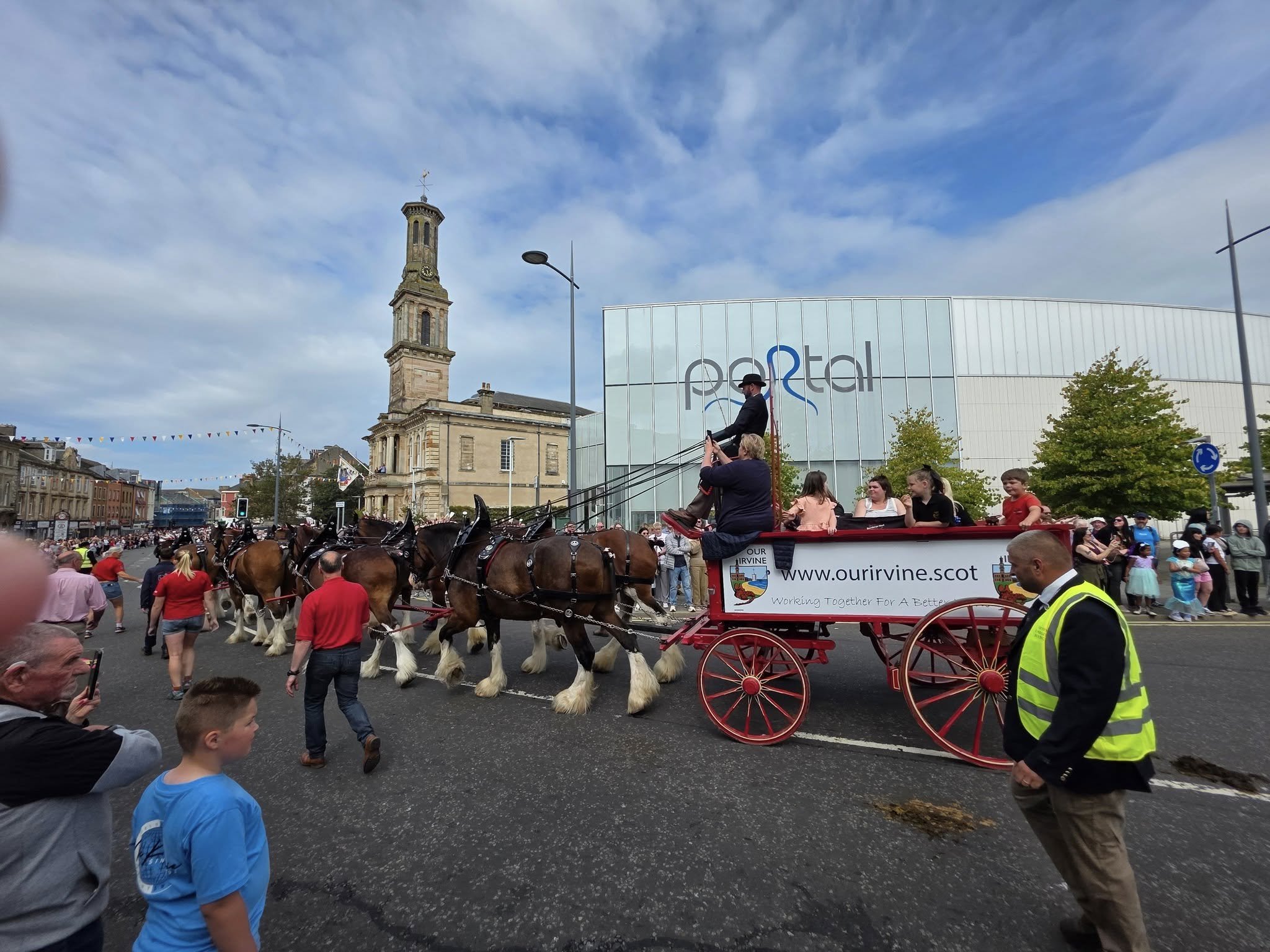 An eight-horse hitch of Clydesdales pulling a red cart branded with the Our Irvine logo down Irvine High Street during the Marymass parade, with the Townhouse clocktower in the background and crowds lining the route.